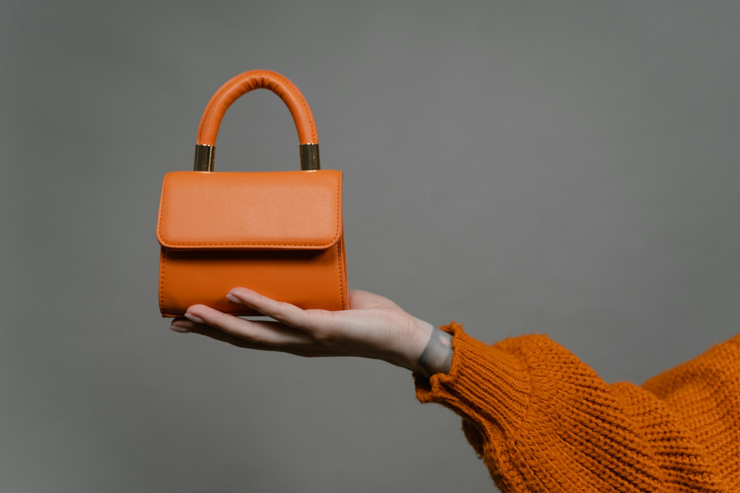 Stylish orange handbag held by a hand against a gray background. Perfect for modern fashion.